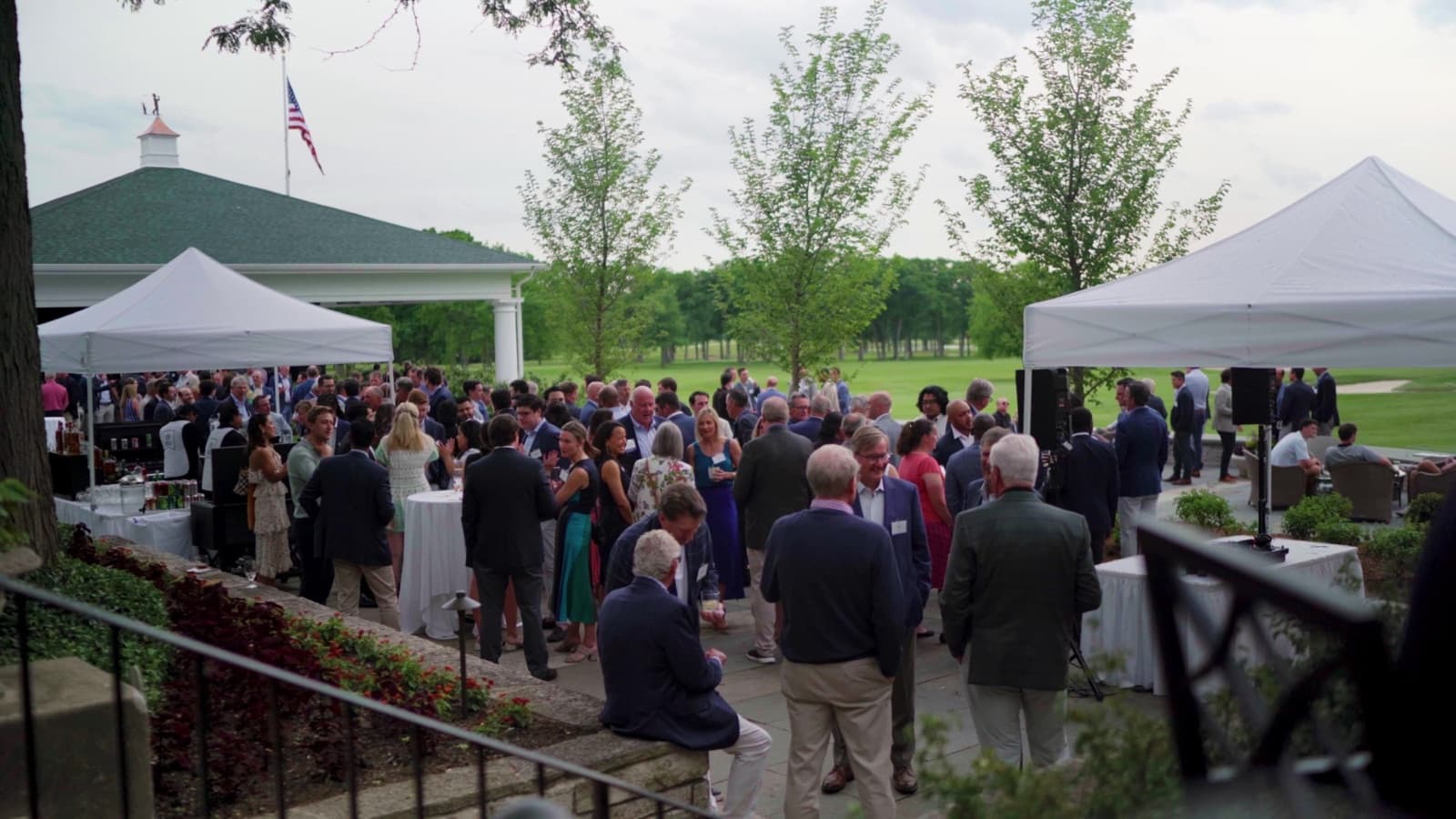 A crowded outdoor donor reception on a country-club lawn with white tents, high-top cocktail tables, and guests in formal attire mingling near a green-roofed pavilion.